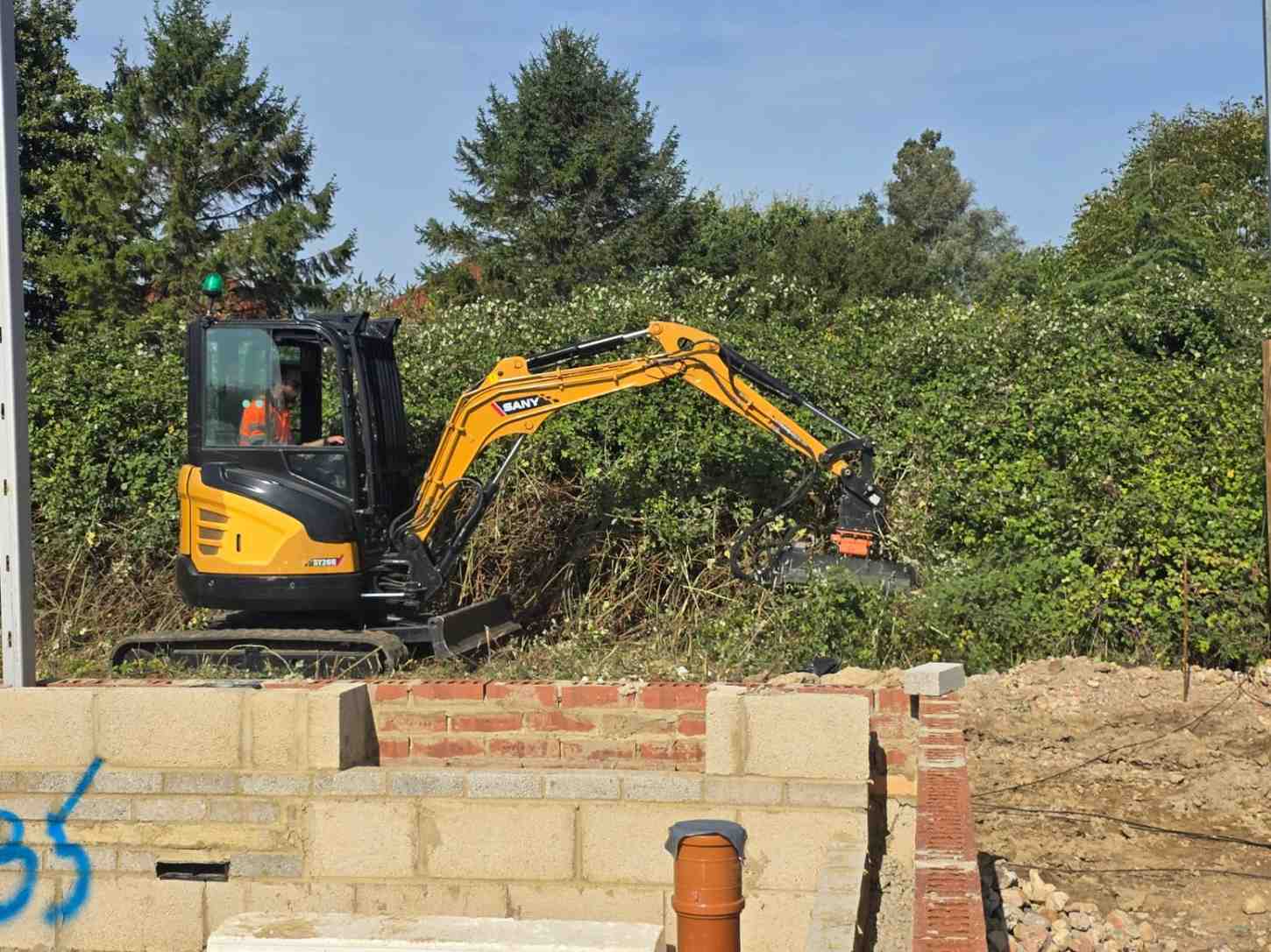 Mini digger clearing overgrown vegetation beside new-build house footings
